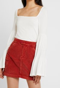 Maglietta bianca a maniche lunghe con scollatura quadrata e maniche svasate abbinata a una mini gonna rossa in velluto a coste dotata di un bottone e tasche laterali.