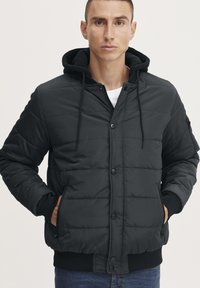 Giacca a vento nera con chiusura a zip e bottoni a pressione, texture trapuntata, cappuccio regolabile e polsini a coste, dotata di tasche laterali e tasca con zip sulla manica.