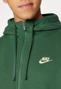 Zelená mikina na zip z měkké látky. Má klokánkovou kapsu, stahovací šňůrky a zlaté logo Nike na levé straně hrudi.
