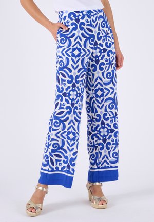 Femme portant un pantalon large avec des motifs géométriques complexes bleus et blancs, associé à des sandales compensées dorées et un haut blanc.