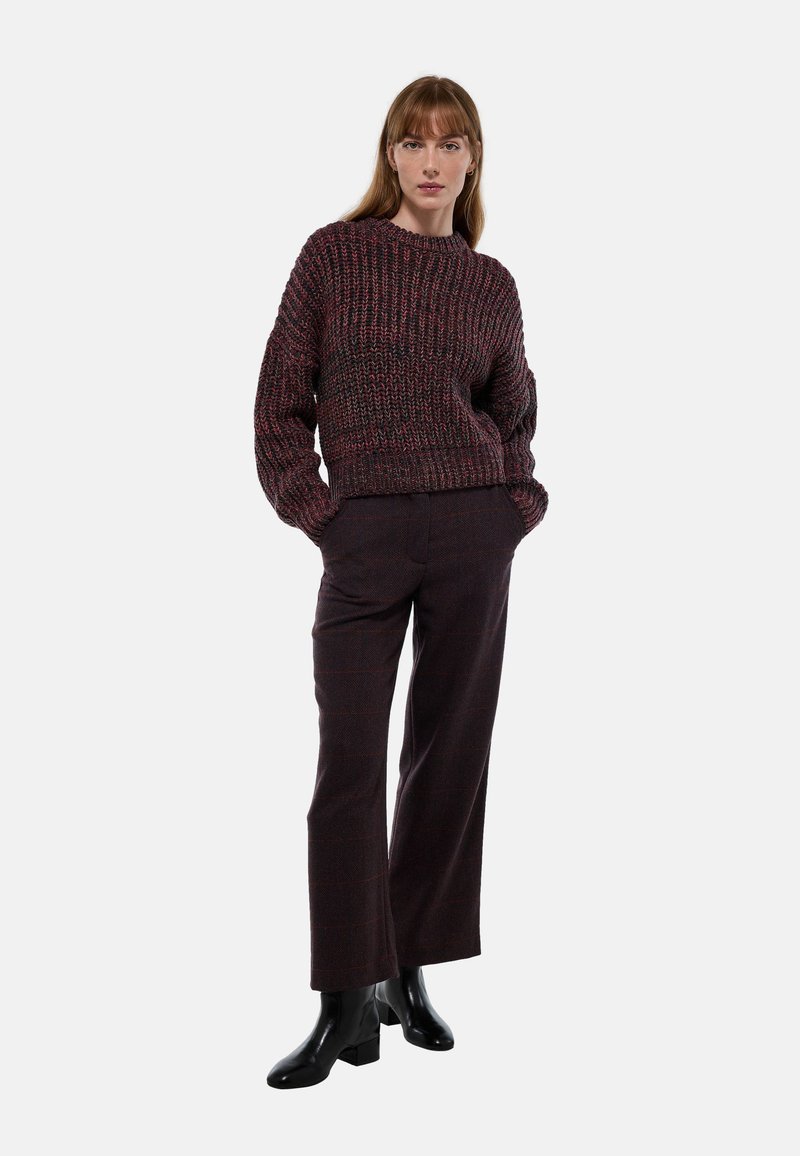 Pull en maille épaisse dans des tons rouge foncé et noir avec un col rond ; associé à un pantalon noir ajusté et des bottines noires brillantes.