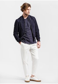 Námornícky zamkový bomber kabát cez tmavé tričko, skombinovaný s bielymi nohavicami a bielymi teniskami s hnedými podrážkami.