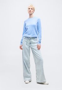 Kobieta stojąca w jasnoniebieskim swetrze, szerokich, wyblakłych dżinsach z sznurkiem oraz białych tenisówkach na jednolitym tle.