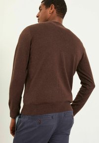 Brauner Strickpullover mit Rundhalsausschnitt, gerippten Bündchen und Saum. Getragen mit navyblauen Hosen, die ein gemustertes Innenfutterdetail zeigen.