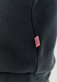 Tmavošedý mikina s rebrovaným lemom a manžetami, so malou ružovou štítkovou značkou na ľavej strane pre branding.