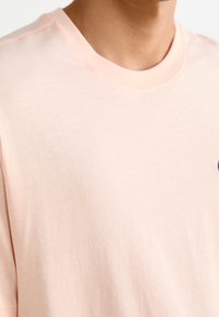 Jasnoróżowa bawełniana koszulka z okrągłym dekoltem, krótkimi rękawami i gładką fakturą. Posiada minimalne szwy i brak widocznych wzorów czy grafik.