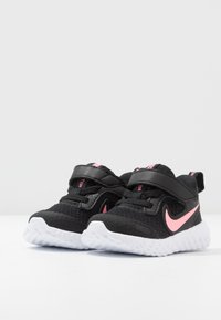 Ein Paar schwarze und rosa Nike-Kinderschuhe mit weißen strukturierten Sohlen und Klettverschlüssen auf einer weißen Oberfläche.