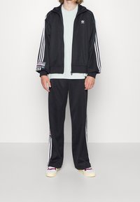 Svart Adidas träningsdräkt med vita ränder på ärmarna och byxorna, med en logopatch på jackan och utsmyckade detaljer på byxorna.