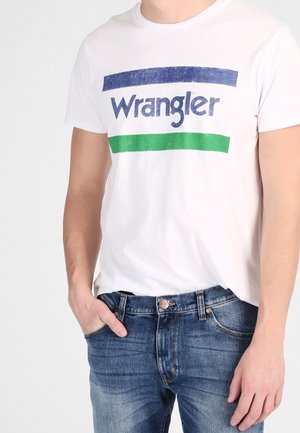 Homme portant un t-shirt Wrangler blanc avec des rayures bleues et vertes, associé à un jean en denim bleu, la main dans la poche.