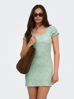 Femme portant une robe courte à manches courtes fleurie vert menthe, des lunettes de soleil foncées et portant un grand sac cabas en daim marron sur fond blanc.