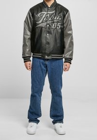 Svart läder och grå ärmar på en varsity-jacka med tryckt logotyp, tryckknappar, randiga ribbade muddar, i kombination med blå jeans och vita sneakers.