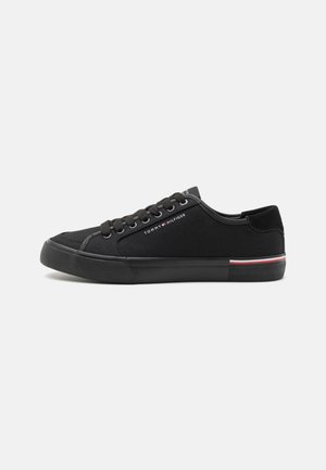 Zapatilla deportiva negra de caña baja con cordones negros, suela negra, pequeño logo de Tommy Hilfiger en el lateral y una franja roja, blanca y azul en el talón.