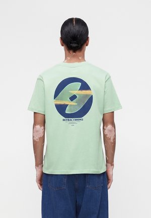 Άτομο με μαλλιά δεμένα προς τα πίσω και λεύκη, που φοράει ανοιχτό πράσινο μπλουζάκι με κυκλικό αφηρημένο σχέδιο και το κείμενο «Refractions» στην πλάτη, σε συνδυασμό με μπλε τζιν.