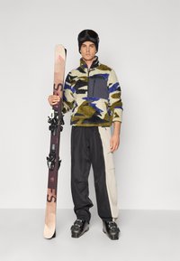 Fleece-Pullover in grün, blau und cremefarbenem Camouflage-Muster, graue Brusttasche; schwarze und beige Skihosen; Skier mit schwarzen Bindungen und Skischuhen.