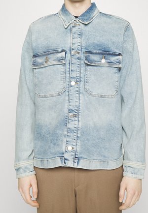 Vaaleansininen farkkutakki, jossa etuosa napitettavissa ja kaksi läppätaskua rinnassa, päällä ruskeiden housujen kanssa.