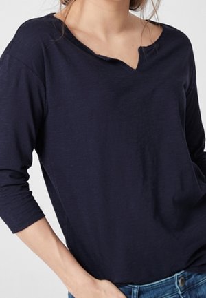 Kobieta w granatowej koszuli z długim rękawem i wycięciem przy dekolcie oraz niebieskich dżinsach, z jedną ręką w kieszeni.