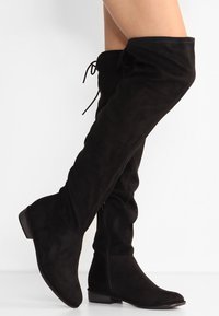 Bottes en suède noires à hauteur de genou avec des petits talons, des fermetures éclair sur le côté et des détails de lacets en haut, portées sur des jambes croisées sur un fond blanc uni.