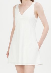 Weißes ärmelloses Kleid mit V-Ausschnitt, A-Linie Silhouette und Taschen, hergestellt aus strukturiertem Stoff, mit minimalistischen Designdetails.