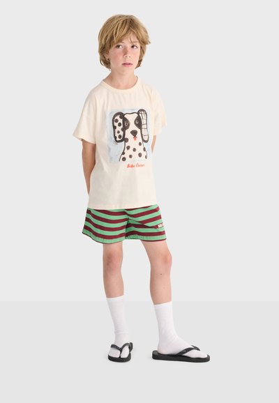 Blond child standing with hands behind back, wearing a white dog-print T-shirt, green and maroon striped shorts, white socks, and black sandals.
