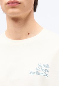 Κοντινό πλάνο ενός ατόμου που φοράει λευκό πουκάμισο με ανοιχτό μπλε κείμενο «No Frills, No Hype, Start Running» στο στήθος και μια μικρή ελιά στον λαιμό.