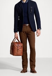 Brun läder weekendväska, marinblå kavaj över en marinblå tröja, bruna byxor och bruna läderskor. Anmärkningsvärt fickdetalj på kavajen.