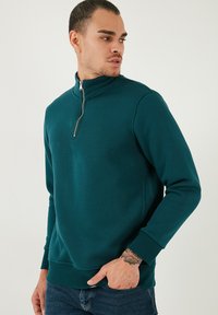 Felpa con zip color verde acqua, realizzata in materiale morbido, con colletto alto e polsini a coste. Il modello la indossa con jeans blu.