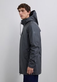 Hombre con una chaqueta impermeable con capucha de color gris oscuro y bolsillos frontales, de pie de lado contra un fondo gris claro liso.