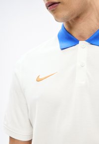 Polo blanc avec col bleu vif et logo Nike Swoosh orange sur la poitrine, porté par une personne dont le bas du visage et le cou sont visibles.