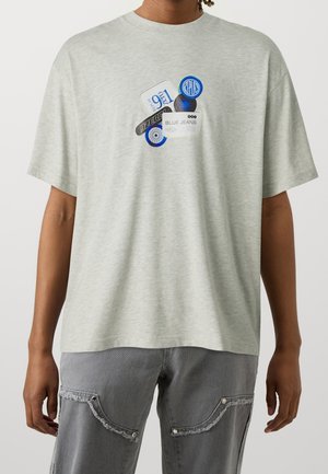 Homme portant un T-shirt gris clair avec des étiquettes graphiques bleues et blanches et un pantalon cargo gris avec des bords de poche effilochés.