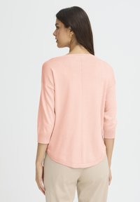 Pull rose clair tricoté à manches trois-quarts avec un ourlet arrondi, mettant en valeur une couture dans le dos et une texture douce.
