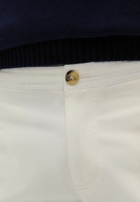 Hvide bukser lavet af bomuld med en glat tekstur. Har en brun knap i taljen og hvid syning. Parret med en marineblå striktrøje.