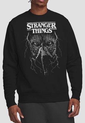 Person wearing black Stranger Things sweatshirt with white graphic of character facing large monster face and lightning bolts.