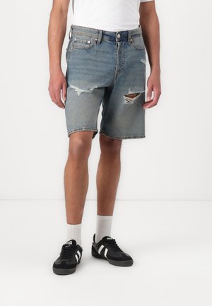 Denim šortsevi svijetloplave boje s izbleđenim naglasima. Imaju dizajn s pet džepova, petlje za remen i ravni kroj. Upareni su s crnim tenisicama.