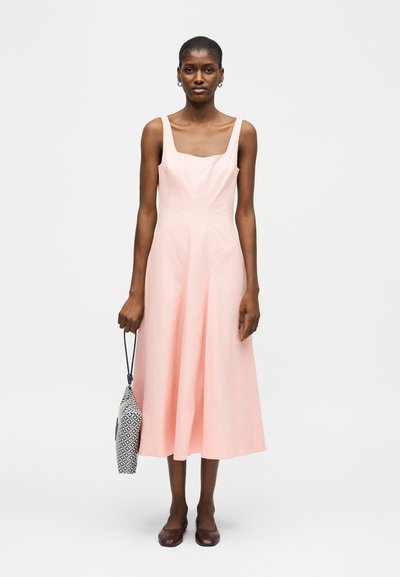 Kvinde iført ærmeløs pastelfarvet lyserød midi-kjole, der holder et mønstret håndtaske i venstre hånd, stående mod en ensartet hvid baggrund.