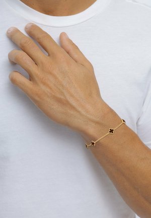 Gold bracelet featuring floral designs with dark accents, linked by delicate chains, worn on a hand against a white shirt background.
