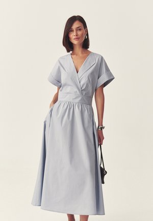 Kobieta o ciemnych, do ramion sięgających włosach, ubrana w jasnoniebieską sukienkę midi z krótkim rękawem i dekoltem w serek, nosząca srebrne kolczyki koła i trzymająca małą czarną torebkę.