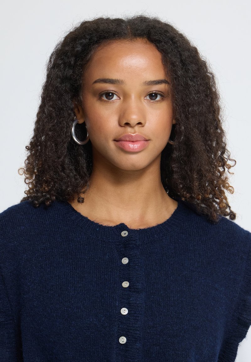 Jeune femme aux cheveux bouclés, portant des boucles d’oreilles en argent et un cardigan bleu marine boutonné, regarde devant elle devant un fond uni clair.
