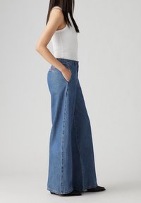 Högmidjade, vida blå jeans i denim, med framfickor, dragkedja och knappstängning, samt en diskret märkeslapp på baksidan.