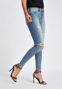 Jeans ajustados de mezclilla azul claro con parches desgastados y rodillas rasgadas, combinados con sandalias de tacón alto negras con una correa de estampado de leopardo.