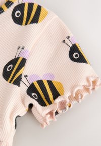 Tissu côtelé rose avec bord volanté et motifs de petites abeilles dessinées en noir, jaune et violet sur une manche de vêtement.