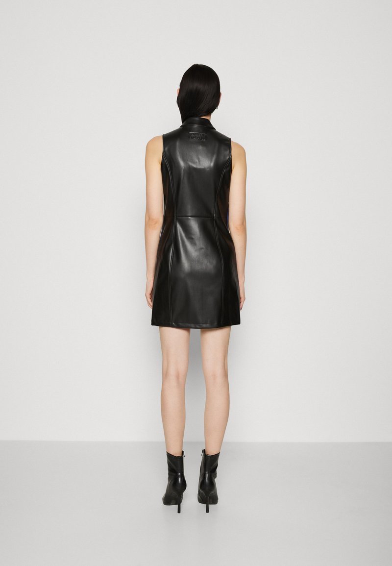 Abito in pelle nera senza maniche con silhouette aderente, dettagli delle cuciture frontali e colletto alto. Abbinato a stivali neri con tacco all'altezza della caviglia.