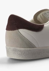 Zapatilla de cuero blanca con acentos de gamuza gris y burdeos. Suela de goma texturizada. Detalles de costura sutiles a lo largo de la parte trasera.