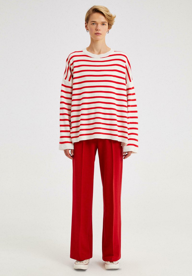 Pulover supradimensionat cu dungi roșii și albe, cu mâneci lungi, asociat cu pantaloni largi roșii. Textură netedă și design casual.