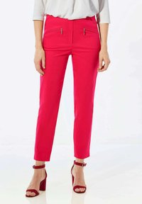Pantalon rose vif à coupe ajustée, avec deux poches avant zippées. Tissu lisse avec une légère brillance. Associé à des escarpins rouges à bride de cheville.