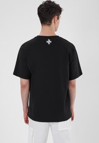 Black cotton t-shirt with a relaxed fit, short sleeves, and a white graphic logo on the upper back. Textured fabric, plain design.