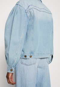 Light blue denim jacket with puffed sleeves, frayed stitching at shoulders, and brass button accents at the cuffs and hem.