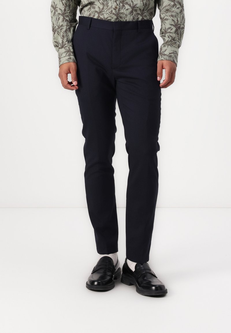 Marineblå slim-fit bukser lavet af glat stof, med en flad frontdesign og bæltestropper. Sammen med sorte loafers og lyse sokker.