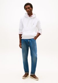 Fehér kapucnis pulóvert rózsaszín ingre húzott férfi kék farmerben és barna tornacipőben, világos háttér előtt áll, egyik kezét a zsebében tartva.