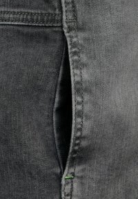 Tela de mezclilla gris oscuro con una ligera textura, costuras visibles y un pequeño detalle verde cerca de la abertura del bolsillo. Corte moderno.