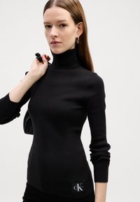 Fekete bordázott garbó pulóver hosszú ujjakkal és testhezálló fazonnal. A csípőn található foltján a Calvin Klein logó látható.